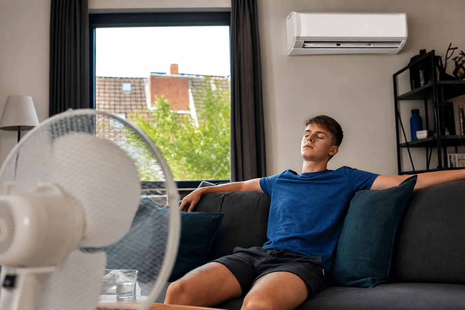 Junge Person sitzt entspannt vor Ventilator im abgedunkelten Wohnzimmer während Sommerhitze